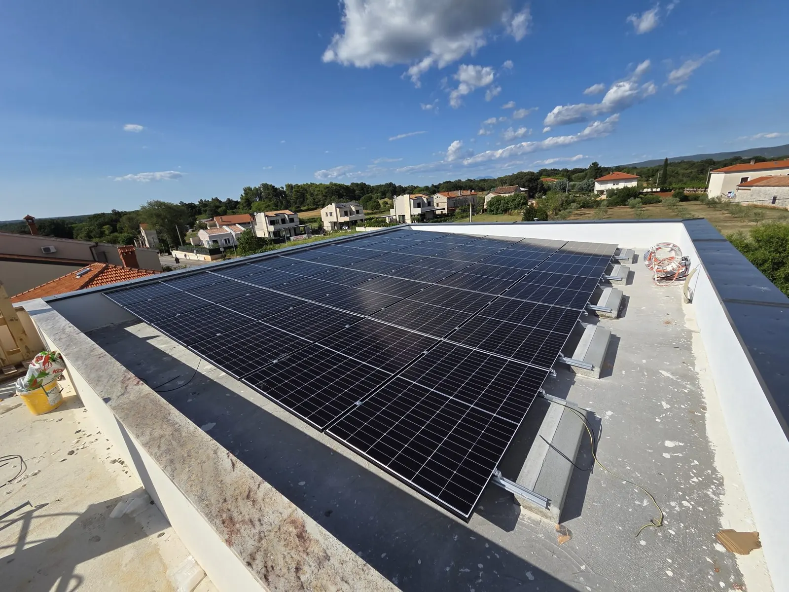 Solarni paneli na ravnom krovu kuće pod plavim nebom, s kućama i zelenilom u pozadini.