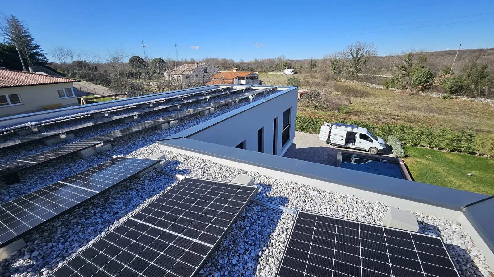 Solarni paneli na ravnom šljunčanom krovu kuće, s poljem i bijelim kombijem u pozadini.